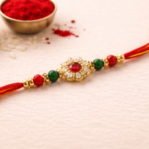 Close-up of floral rakhi with red center stone, pearl detailing, and red-green beads on traditional red-yellow thread.